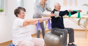 Elderly Man And Woman Exercising On Gymnastic Balls During Physiotherapy Session At Hospital